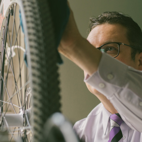 A business man wearing glasses, inspecting the tyre on a bicycle.