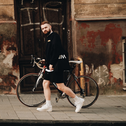 A bearded young man, walking a bicycle along past a dark, graffitied wall, looking at the camera in annoyance.