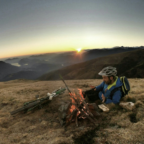 A mountain biker lying down on a hillside, warming his hands on a small fire he has made. His bicycle is on the grass opposite him, a sunset in the distance.