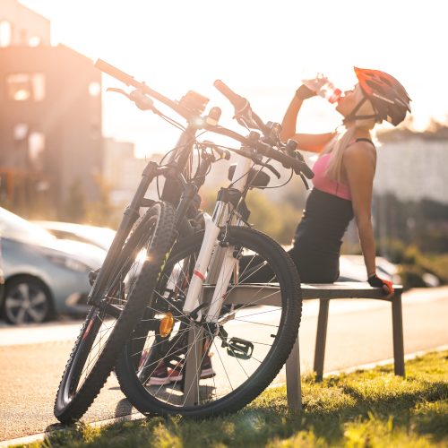 A woman in cycling gear, sitting on a bench next to two mountain bikes and swigging from a drink bottle.