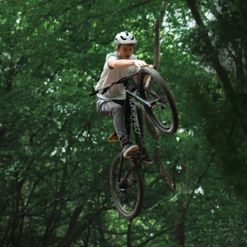 A young mountain biker doing a jump in the forest, touching his front tyre while he's in the air.