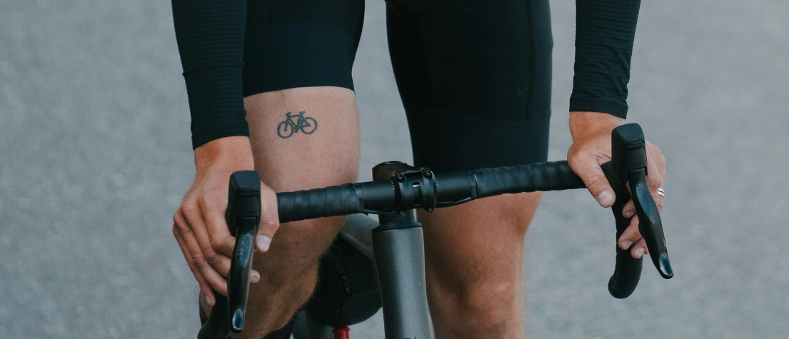 Close-up of the bicycle tattoo on a bike rider's thigh. They are wearing black lycra and riding a drop-bar bike.