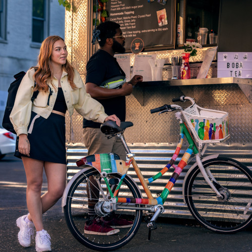 A young woman stands next to a colour-wrapped bicycle, at a boba tea outdoor stall. A man behind her is ordering from the stall.