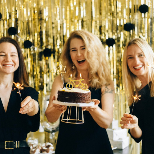 Three women wearing black and standing against a gold streamer curtain. The middle woman holds a cake with a bicycle-shaped candle on it, while the other two hold sparklers.