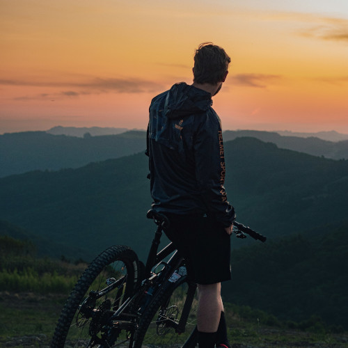 A man leans against a mountain bike, looking at a beautiful sunset over the mountains in the distance.