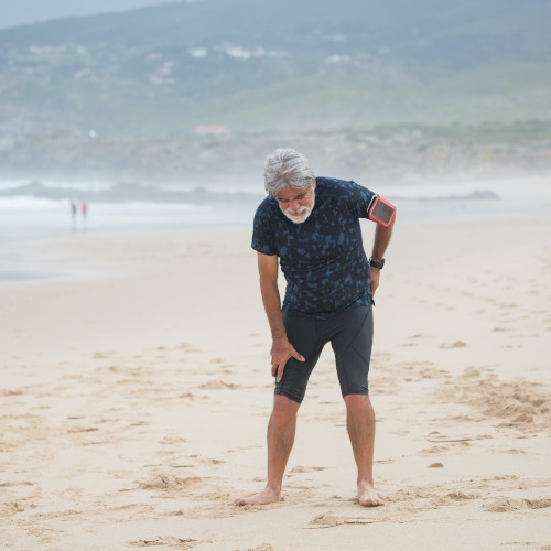 An old man on a beach, bending over and holding his back, grimacing in pain.