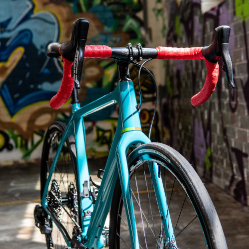 A turquoise and red Open UP bicycle, custom-built by Bio-Mechanics Cycles & Repairs. It's photographed in a car park against a colourful graffitied backdrop.