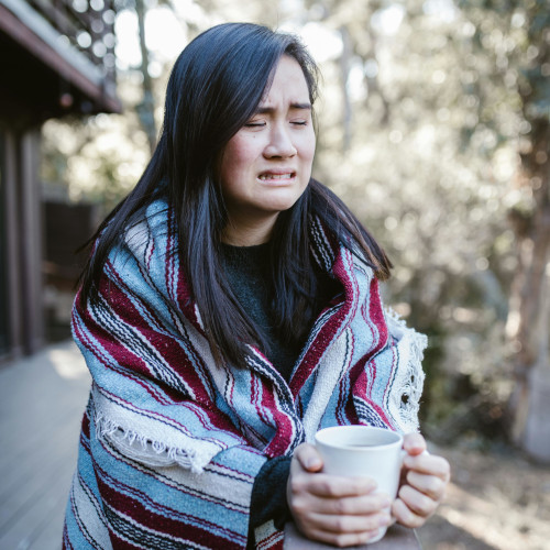 A woman wrapped in a blanket, standing on the deck of a timber house and crying. She's holding a mug.