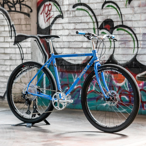 A Surly Midnight Special bicycle, custom-built by Bio-Mechanics Cycles & Repairs. It's photographed in a car park against a colourful graffitied backdrop.