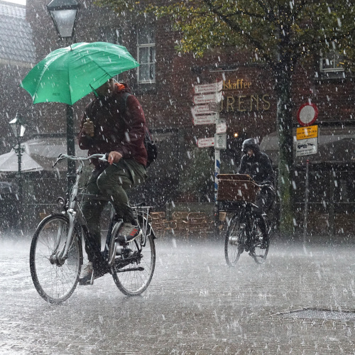 Cyclists riding through a city during a downpour. The leading rider is holding a green umbrella over themselves.