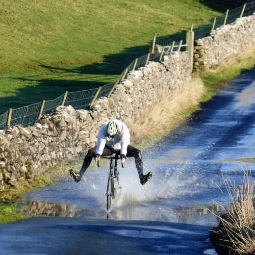 A man cycling through a puddle in a dip in a country road, lifting his feet to avoid getting splashed.
