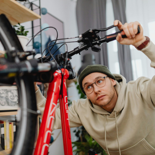 A young man with glasses on, balancing a bicycle on its rear wheel and examining the cockpit. We appear to be in his living room.