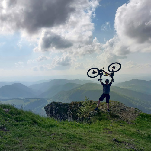 A mountain biker standing on a cliff, holding his bike above his head and looking out at the mountains beyond.