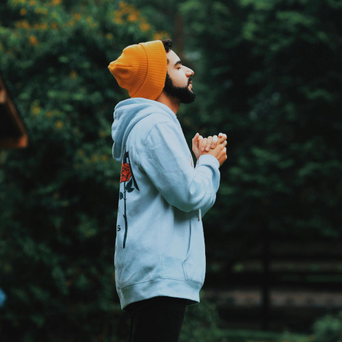 A bearded man standing outdoors, hands clasped, praying.