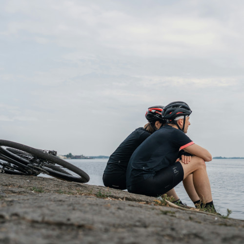 A male and female cyclist sit by a body of water, looking to the horizon, their bikes lying down behind them.