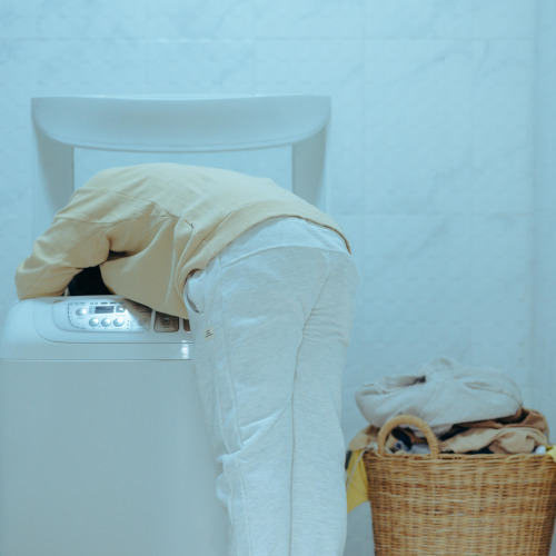 A person leaning far over to look inside a top-loading washing machine.