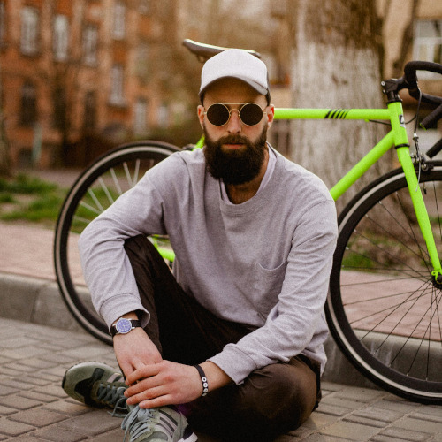 A bearded hipster sitting on the ground in front of his bike.